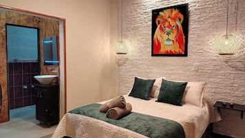 Une chambre à coucher avec un lit, des oreillers, un tableau accroché au mur, et une salle de bain avec un lavabo et un miroir.
