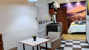 Quarto compacto com cama, uma mesinha com planta, cozinha compacta e um mural da paisagem urbana.