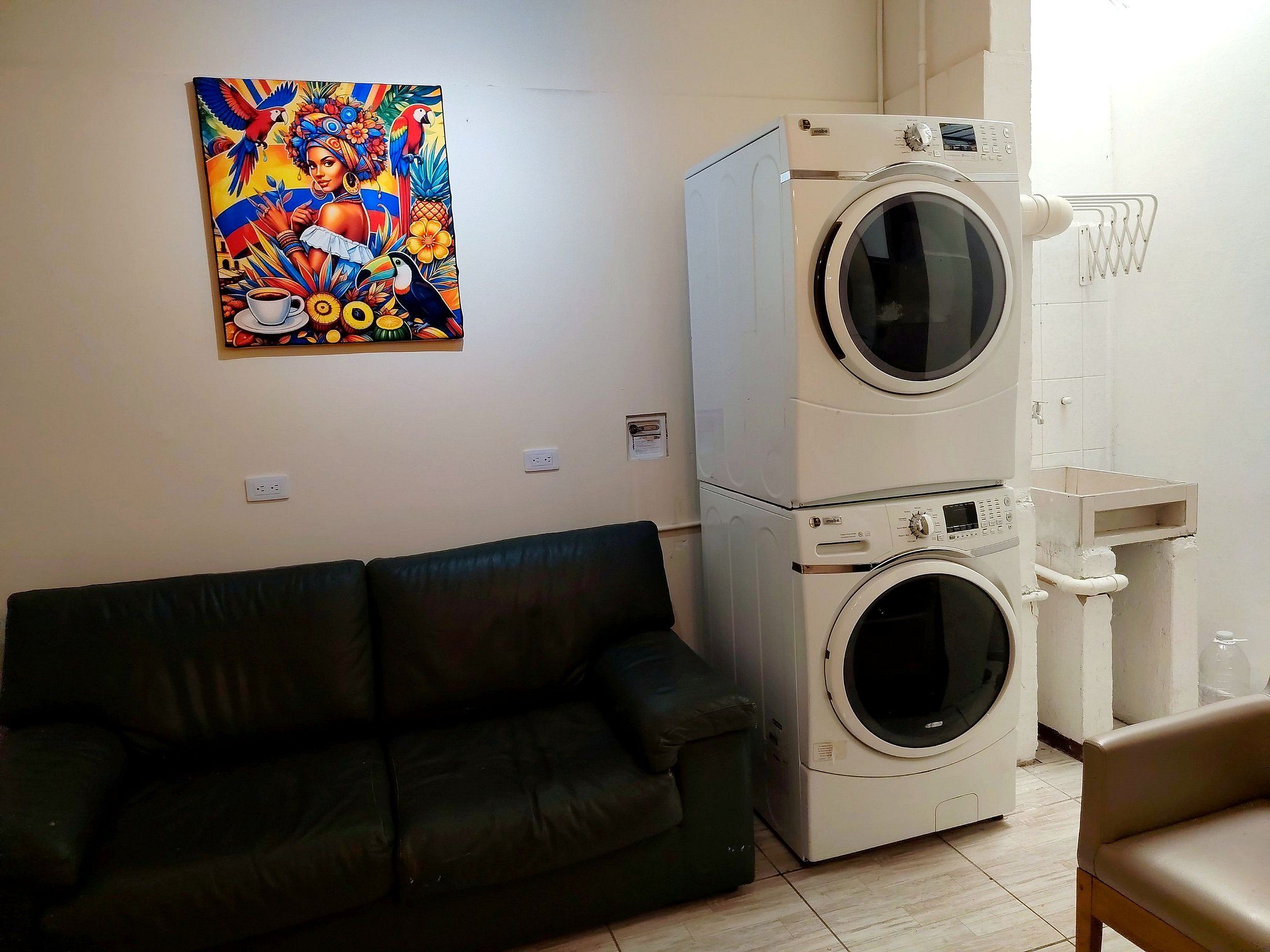 Una habitación con un sofá de cuero negro, lavadora y secadora apiladas, y un cuadro de colores en la pared.
