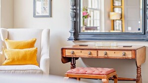Une pièce avec un canapé blanc, des coussins jaunes, un bureau en bois doté de tiroirs, un petit tabouret et un grand miroir qui reflète un mur jaune et une fenêtre.