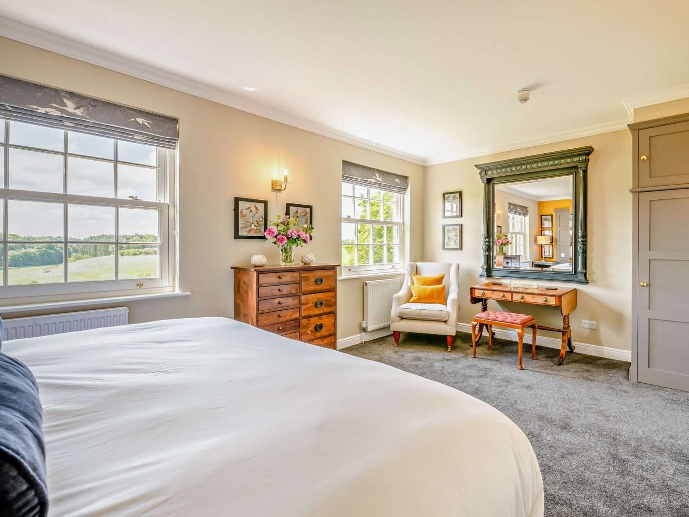 A bedroom with a large bed, a dresser, a chair, a desk, and a mirror. There are windows with views of greenery.