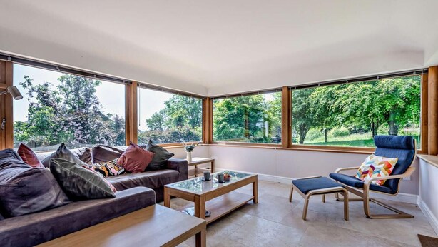 A modern living room with a sofa, a coffee table, and a chair, large windows offering a view of trees and greenery. - 3 Bed in Carmarthen (oc-92116) (Gwernogle)