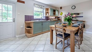 A modern kitchen with a dining area, featuring a wooden table and chairs, a built-in oven, and a window providing natural light. - 3 Bed in Carmarthen (oc-92116) (Gwernogle)