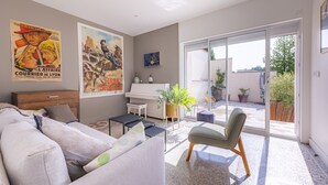 Sala de estar con sofá, piano, mesa de centro y una silla. Hay dos carteles en la pared, una planta y un ventanal corredizo que da a un espacio exterior con plantas en macetas.
