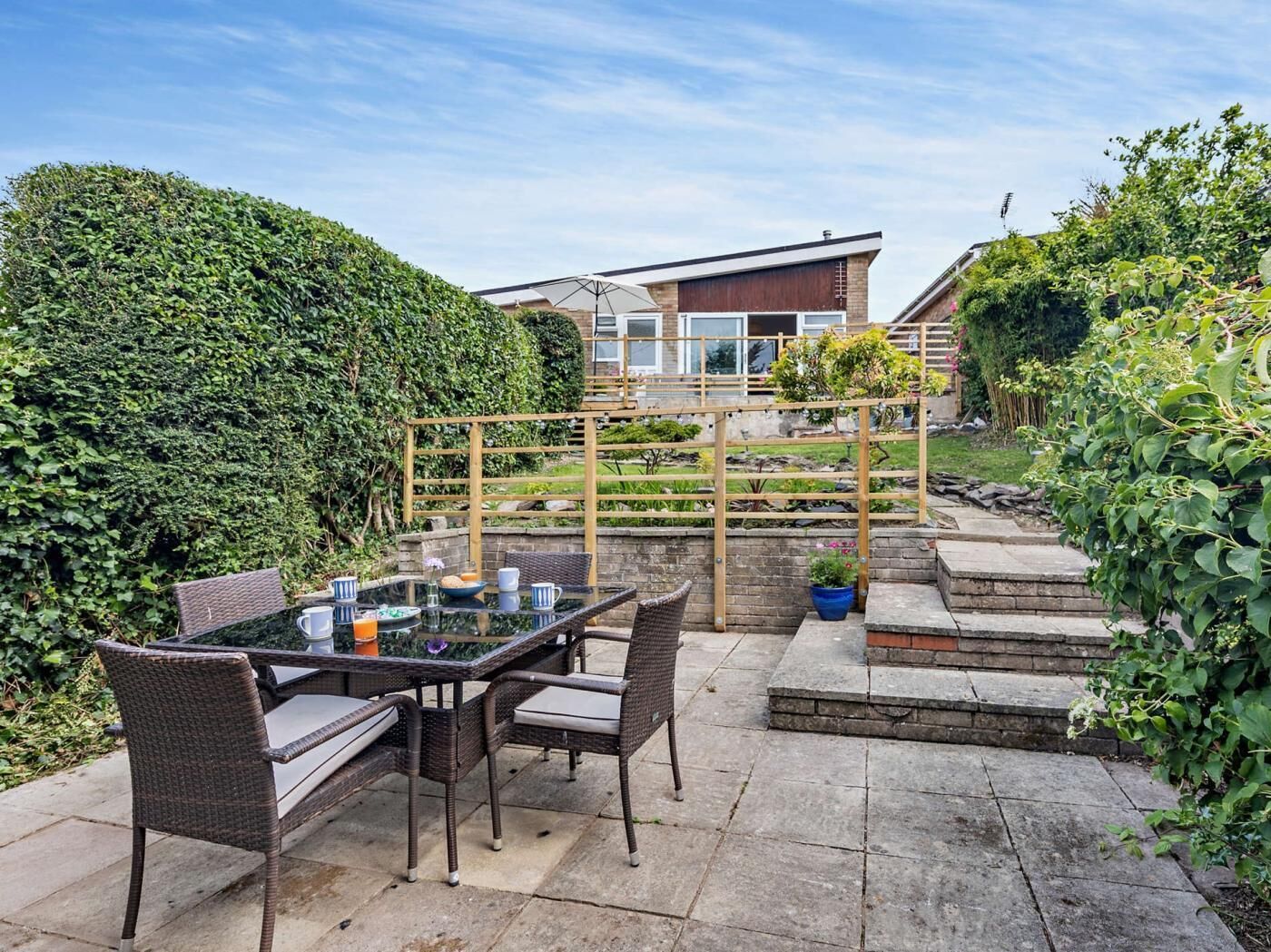A patio with a glass table and wicker chairs, a wooden pergola, and a hedge.