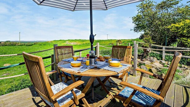 Eine Holzterrasse mit rundem Tisch, Stühlen und einem Sonnenschirm, mit Blick auf eine grüne Landschaft.