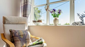 Ein Zimmer mit Fenster, ein Sessel mit Kissen, eine Vase mit Blumen und eine Hirschskulptur.