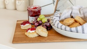 Eine Holzplatte mit Scones, einer Schüssel Trauben und einem Marmeladenglas.
