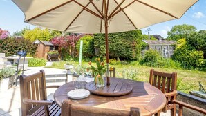Une table ronde en bois, surmontée d’un parasol beige, entourée de chaises en bois, installée à l’extérieur avec vue sur le jardin.