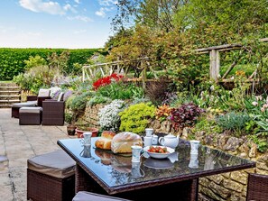 Une terrasse extérieure aménagée avec des meubles en osier, une table en verre et une variété de plantes et de fleurs.