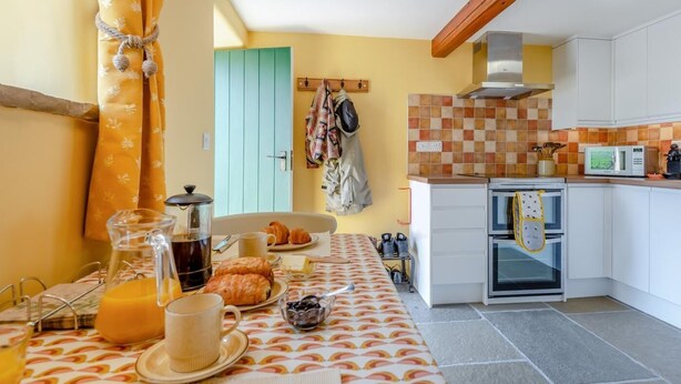 Eine Küche mit Frühstücksangebot, darunter ein Tisch mit gemustertem Tischläufer, eine Kaffeekanne, Croissants und ein Krug Orangensaft.