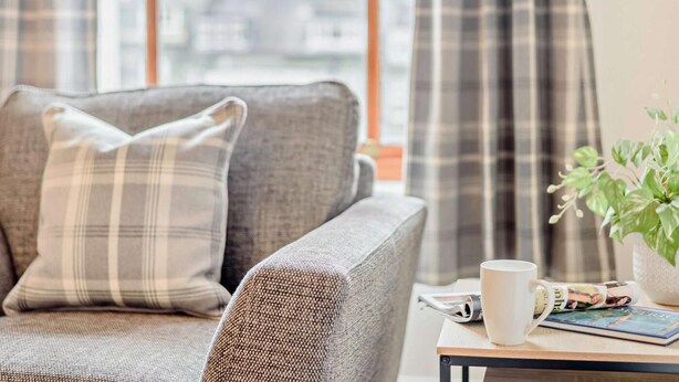 Ein graues Sofa mit einem karierten Kissen, einer weißen Tasse, einer Pflanze und einer Zeitschrift auf einem kleinen Tisch.