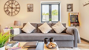 Ein Wohnzimmer mit grauem Sofa, einer großen Wanduhr, einem Fenster mit Blick, einer Pac-Man Spielmaschine und einem kleinen Tisch mit Büchern und Keksen.