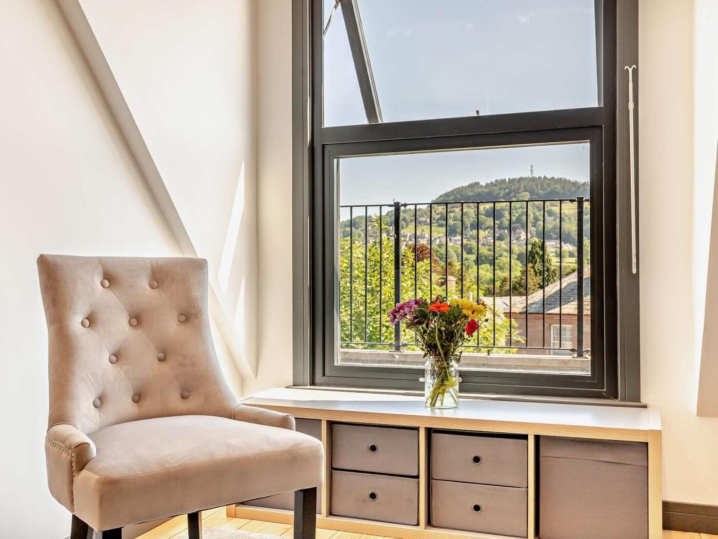 Una habitación con ventana, una silla, un florero con flores y un banco de madera con cajones.