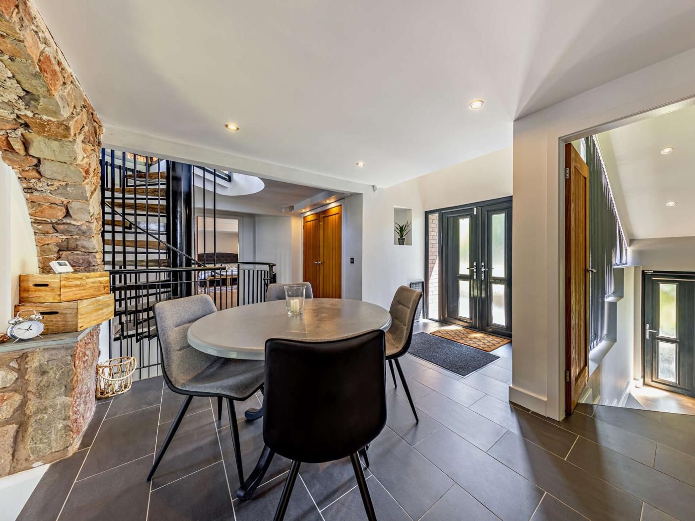 A modern interior with a dining area featuring a round table and chairs, a staircase with a metal railing, and a stone wall.