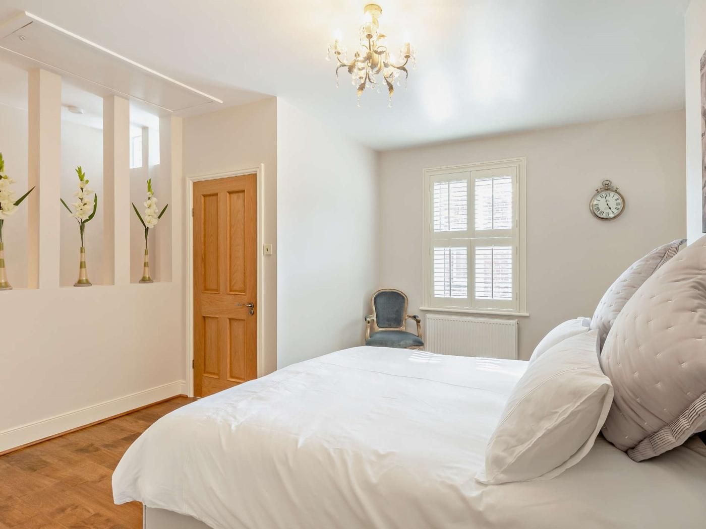 A bedroom with a large bed, a wooden door, a window with blinds, a chair, and a wall clock.