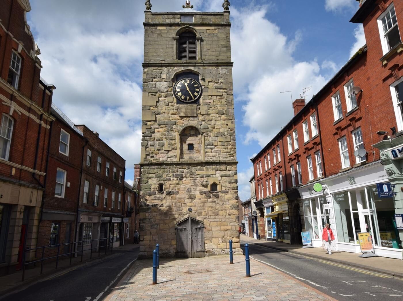 A stone clock tower with a clock face, surrounded by brick buildings and a paved street.
