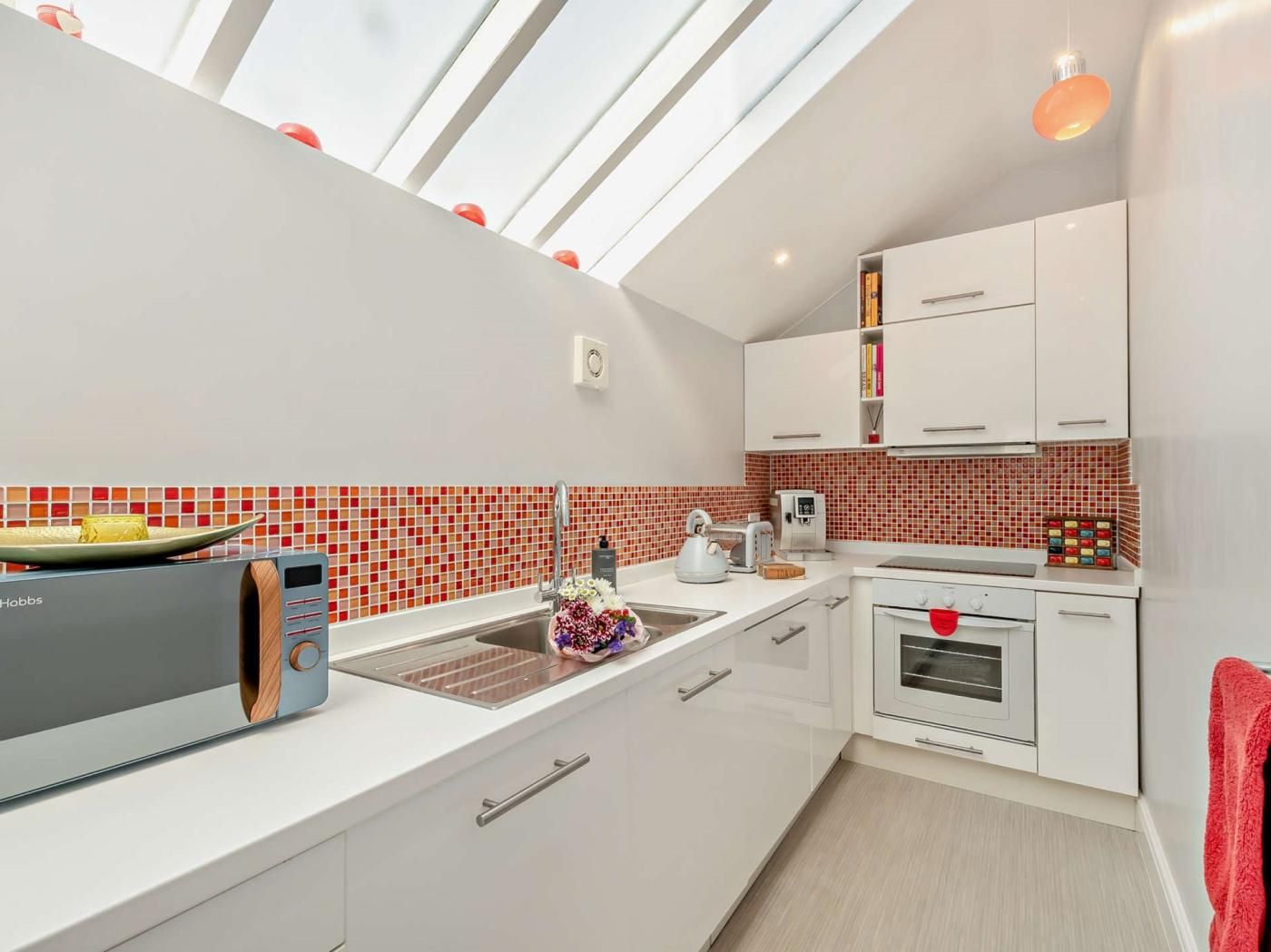 A modern kitchen with a skylight, white cabinets, a red-tiled backsplash, and a red towel hanging on the oven door.