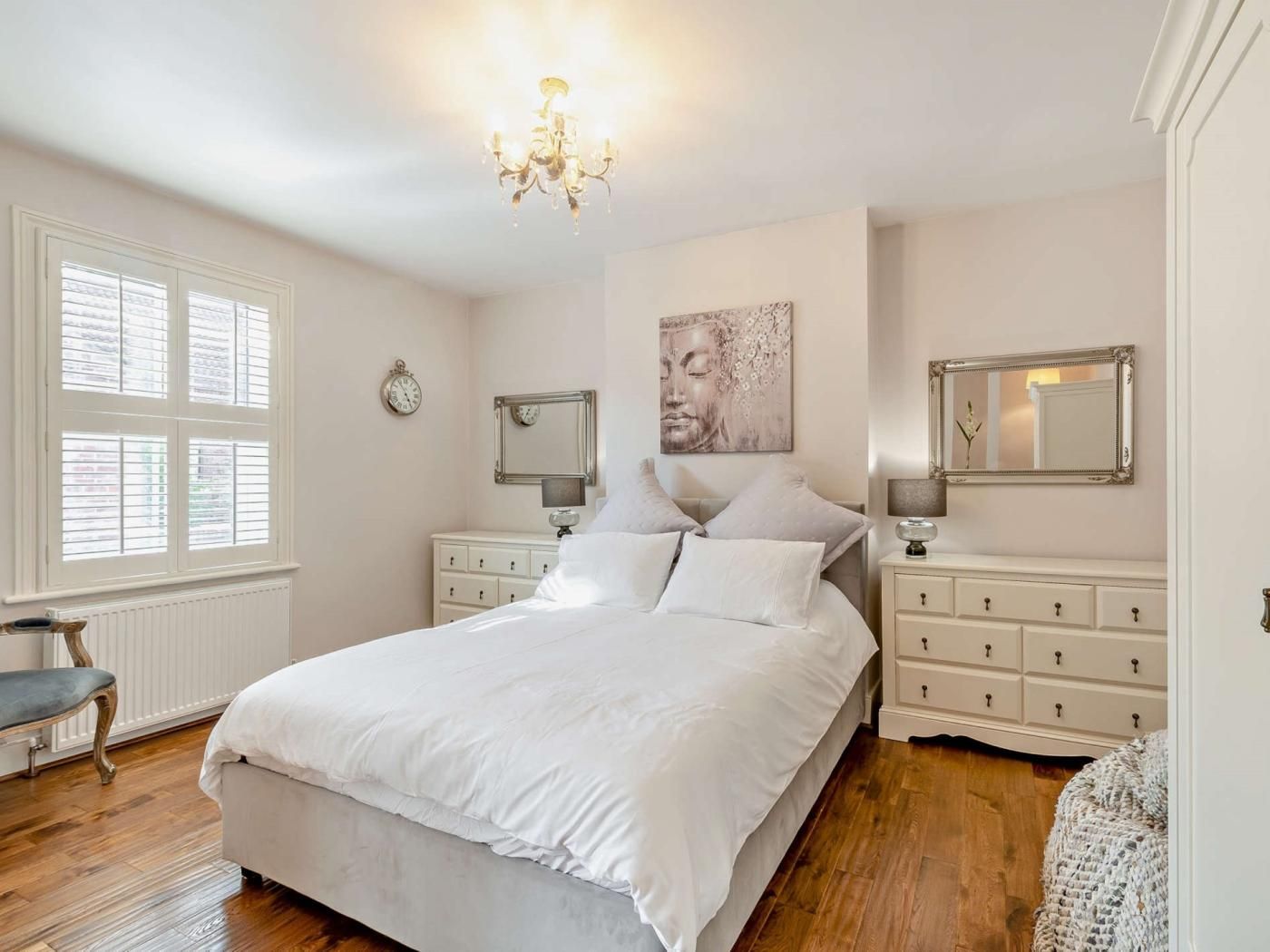 A bedroom with a bed, bedside tables, a chandelier, a window with blinds, and a wall clock.