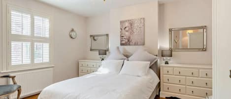 A bedroom with a bed, bedside tables, a chandelier, a window with blinds, and a wall clock.