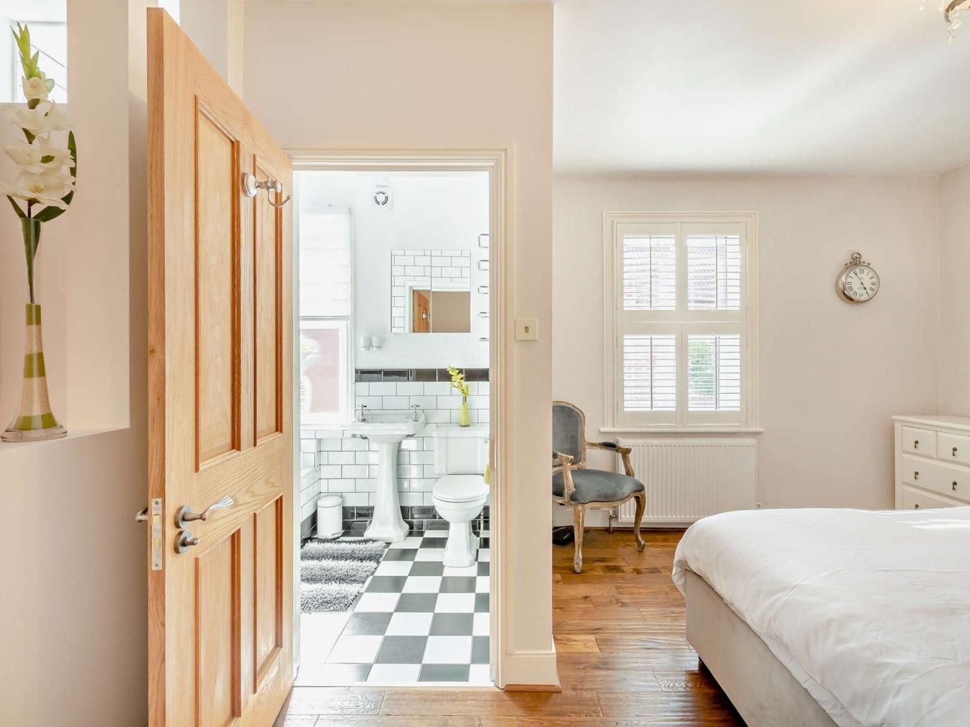 Un dormitorio con una cama, una silla, un tocador y vistas a un baño con lavamanos e inodoro.