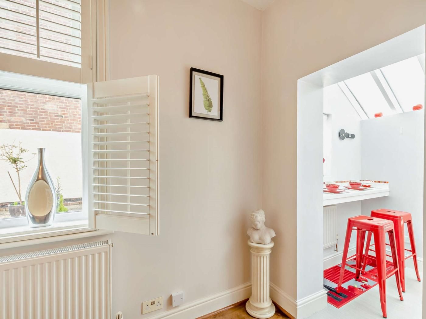 Una habitación con un radiador, una ventana con persianas, un cuadro en la pared y un taburete rojo.