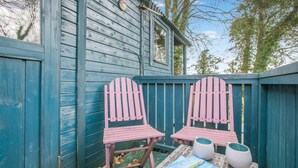 A wooden deck with two pink Adirondack chairs, a table with magazines, and a blue wooden structure. - 1 Bed in Launceston (oc-91964) (Tregadillett)