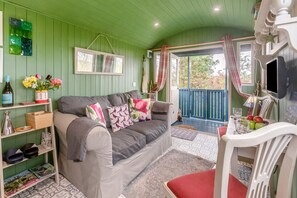 A cozy room with a green wall, a grey sofa with floral cushions, a wooden side table, and a dining area with a white chair and table. - 1 Bed in Launceston (oc-91964) (Tregadillett)