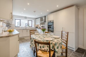 A modern kitchen with white cabinetry, a dining table set for four, and a large window providing natural light. - 3 Bed in Grange-over-sands (oc-92389) (Upper Allithwaite)