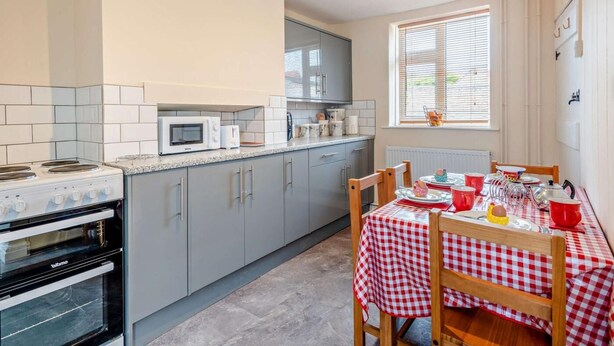 A compact kitchen with a stove, microwave, and oven, a dining table with a checkered tablecloth, and a window with blinds. - 2 Bed in Fakenham (oc-92173) (Fakenham)