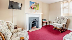 A living room with a fireplace, floral armchairs, a television, and a bookshelf. - 2 Bed in Fakenham (oc-92173) (Fakenham)