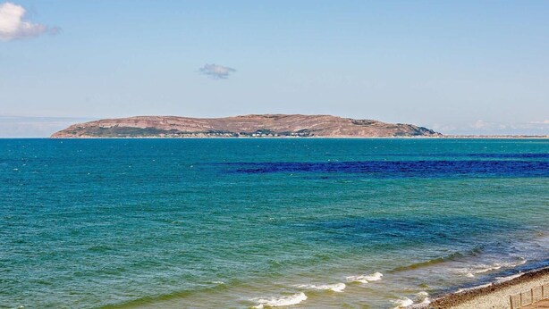 Vista costera con aguas azul turquesa, una playa de arena y una isla rocosa en la distancia.