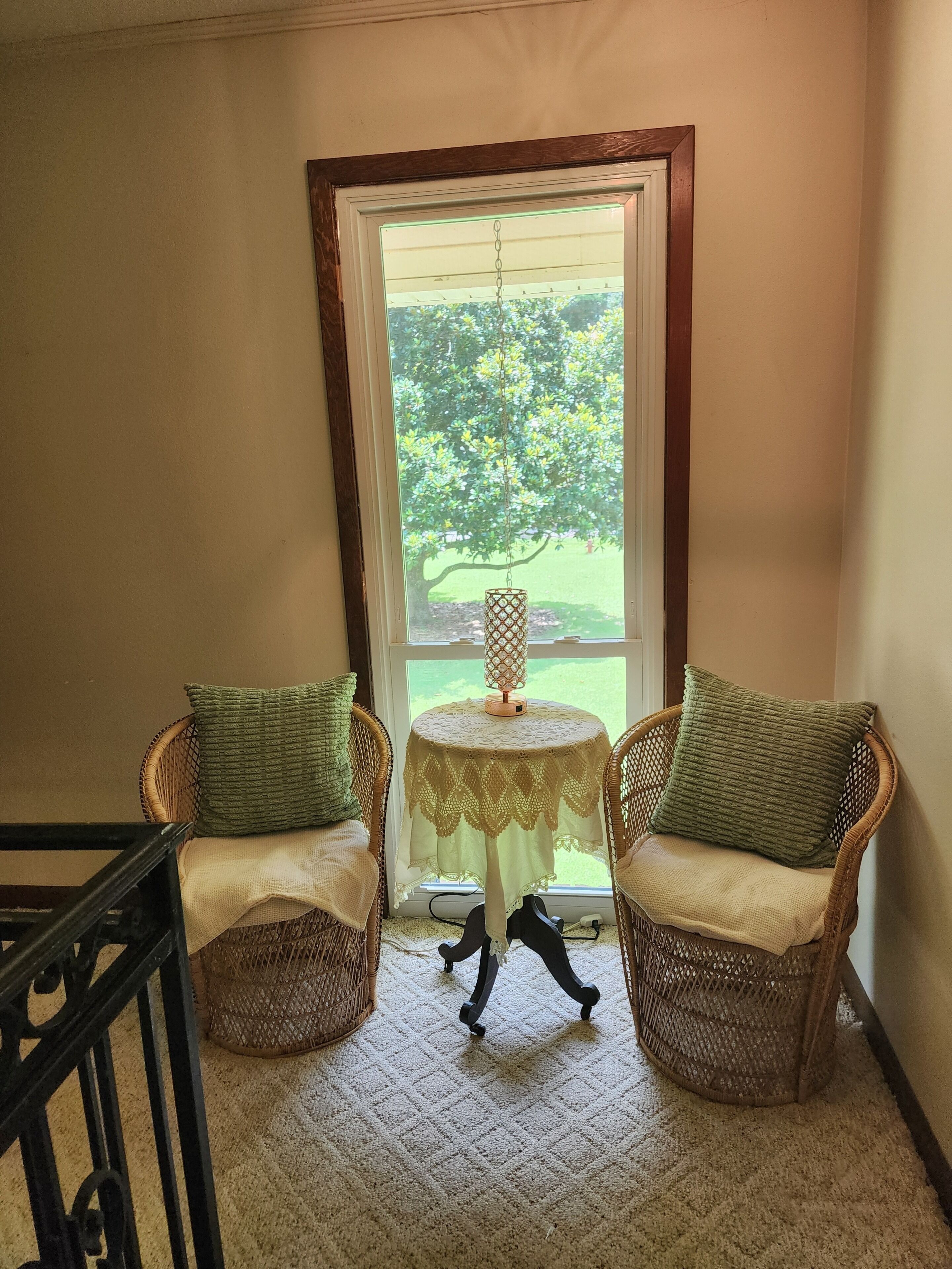 A corner room with two wicker chairs, a small table with a lace tablecloth, and a view of a tree through a window.