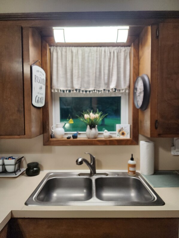 A kitchen with a double sink, a window with a view, a clock, and a sign on the wall.