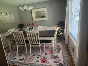 A dining area with a table, chairs, a floral rug, a ceiling light, a mirror, and a window with blinds.