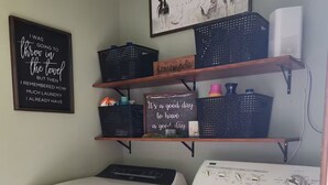 A laundry area with a front-loading washer and dryer, shelves with baskets and decorative signs, and a framed picture on the wall. - The Madison
2 story colonial style home built in 1978, with my father's own hand (Crossett)