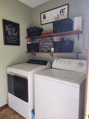 Un área de lavandería con lavadora y secadora de carga frontal, repisas con cestos y letreros decorativos, y un cuadro en la pared.
