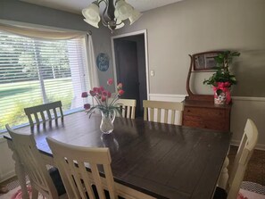 Comedor con mesa de madera oscura, sillas blancas, un florero con flores, una planta en maceta y una araña.
