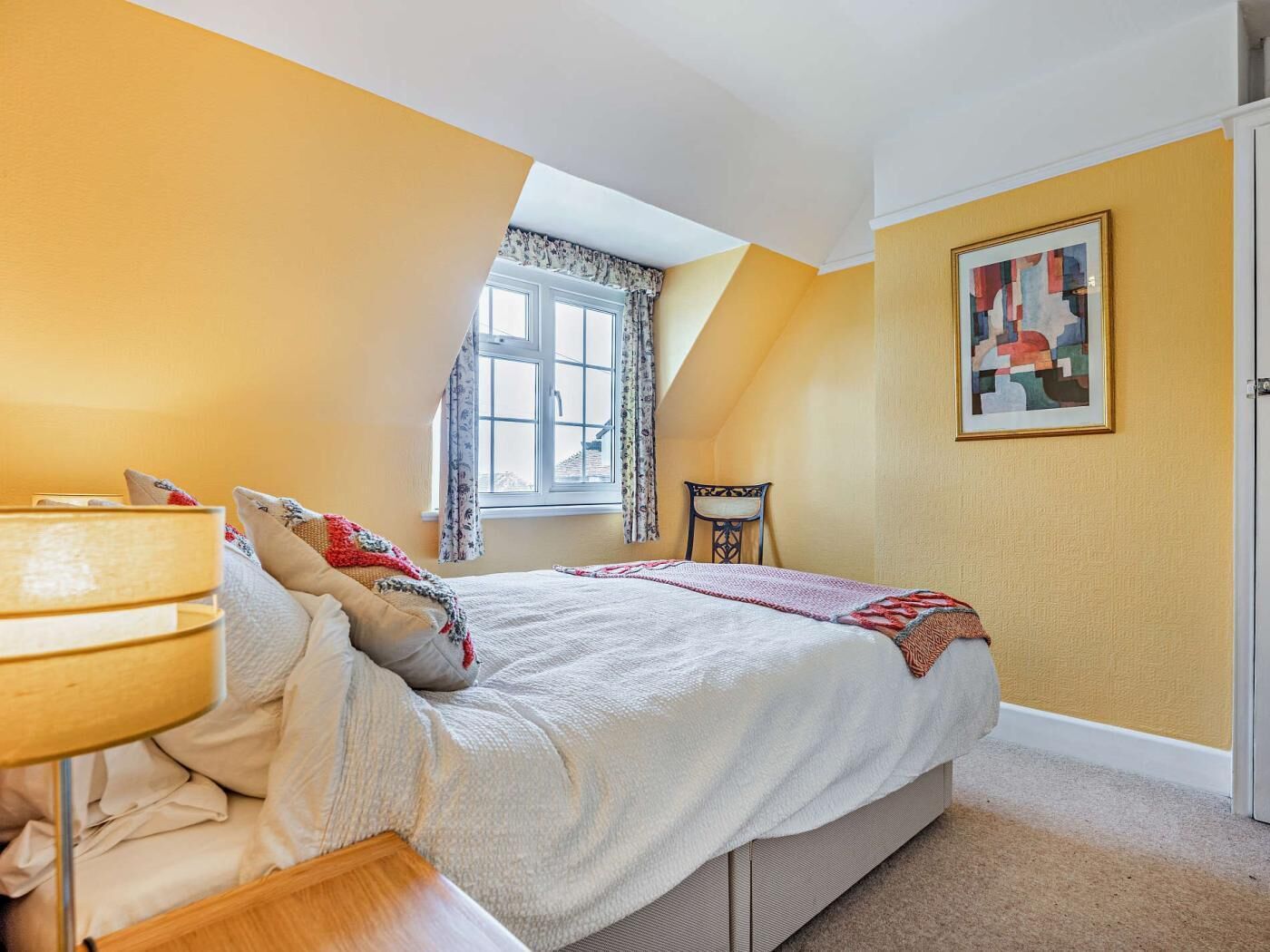 Une chambre à coucher avec un lit, une chaise, une lampe, une fenêtre et un tableau au mur.