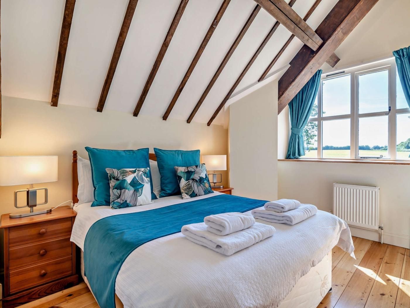 Un dormitorio con techo inclinado, cama con ropa de cama azul y blanca, dos lámparas de noche, una mesita de noche de madera y una ventana con cortinas.
