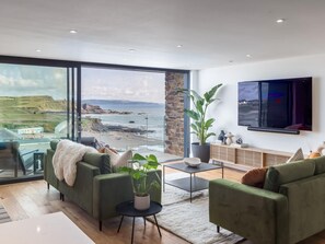 A modern living room with a large window overlooking a coastal landscape, a flat-screen TV mounted on the wall, and a minimalist coffee table. - 2 Bed in Bude (oc-92484) (Bude)
