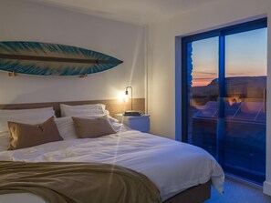 A hotel room with a bed, a nightstand, a wall-mounted surfboard, and a large window with a view of a mountainous landscape. - 2 Bed in Bude (oc-92484) (Bude)