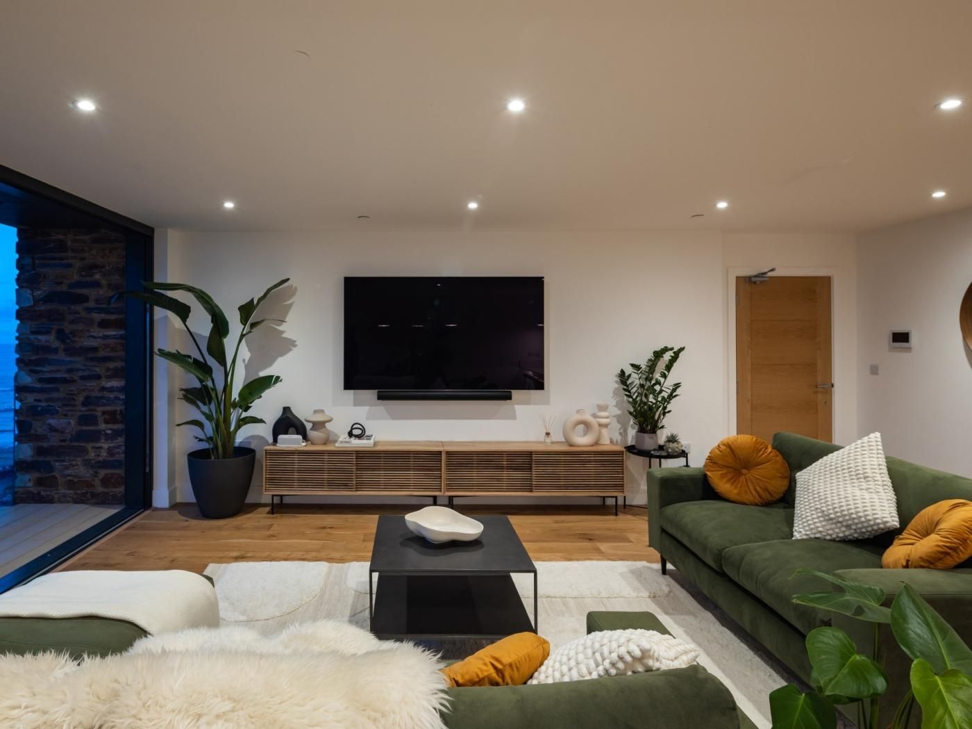 A modern living room with a flat-screen TV, a green sofa, a white furry rug, and a wooden bench.