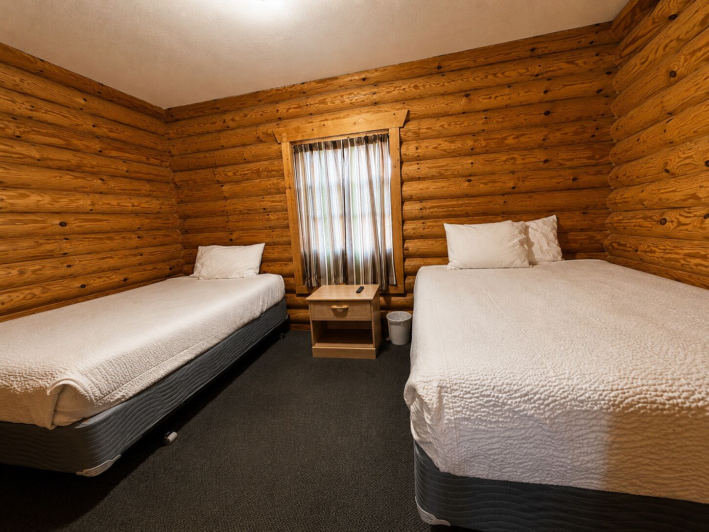 Habitación tipo cabaña con dos camas, una mesita de noche de madera, una ventana con cortinas y piso alfombrado.