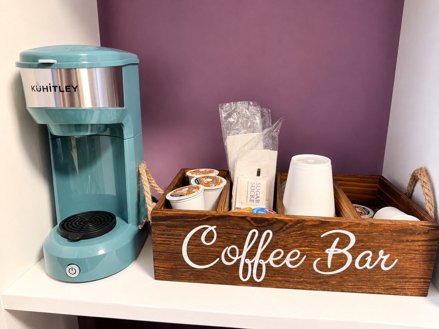 Una cafetera, sobres de azúcar, una taza de café y una caja de madera con la etiqueta 'Coffee Bar' en un estante.