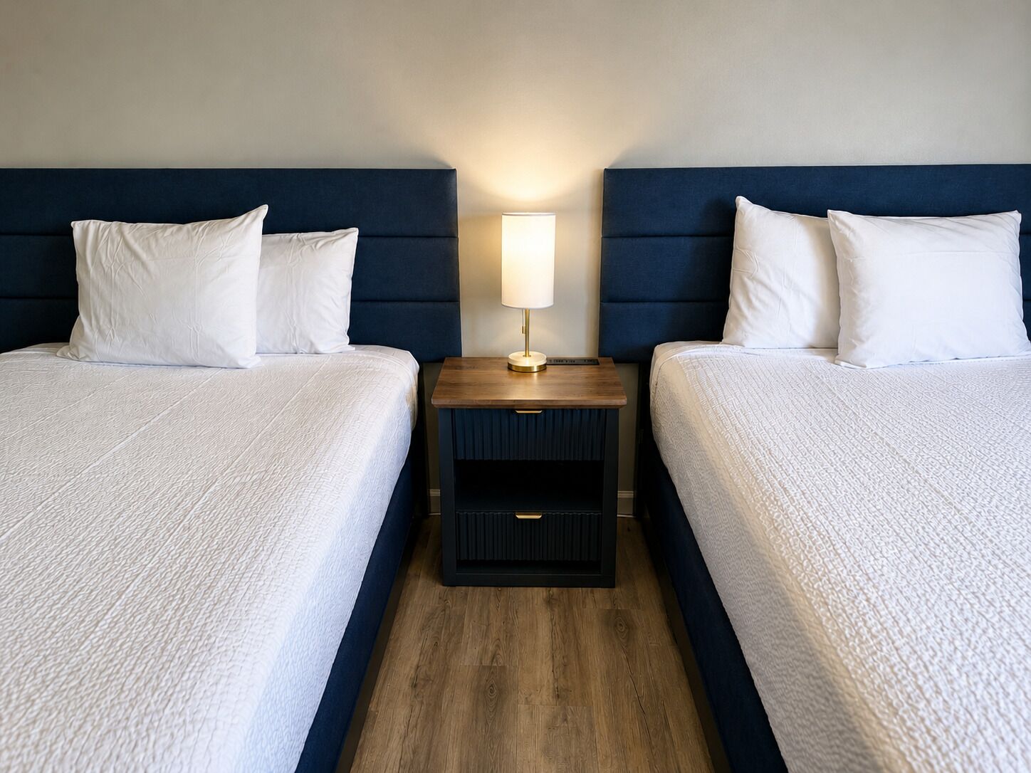 Habitación de hotel con dos camas, una mesita de noche con lámpara y suelo de madera.