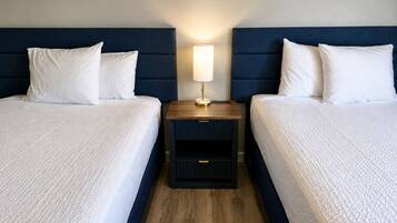 Habitación de hotel con dos camas, una mesita de noche con lámpara y suelo de madera.