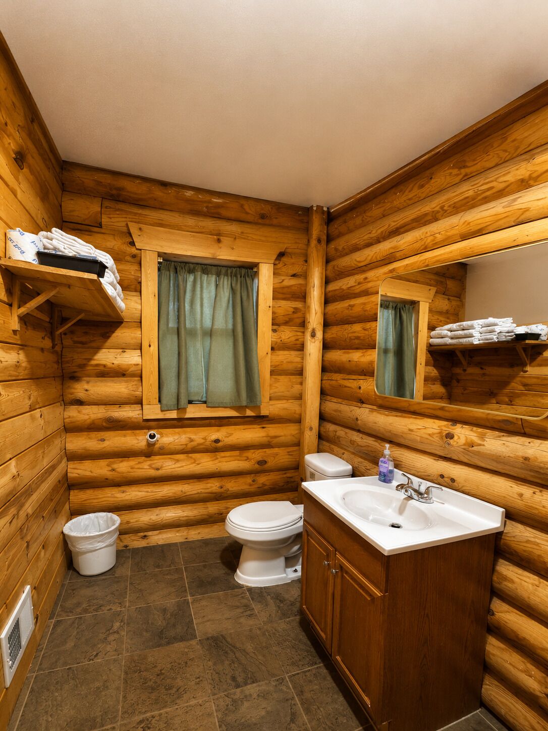 A wooden cabin bathroom with a toilet, sink, and a window with curtains.