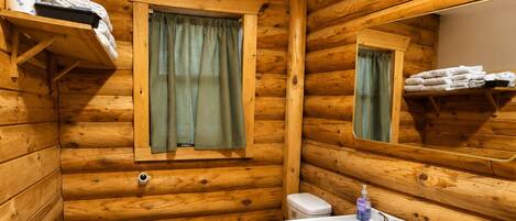 A wooden cabin bathroom with a toilet, sink, and a window with curtains.