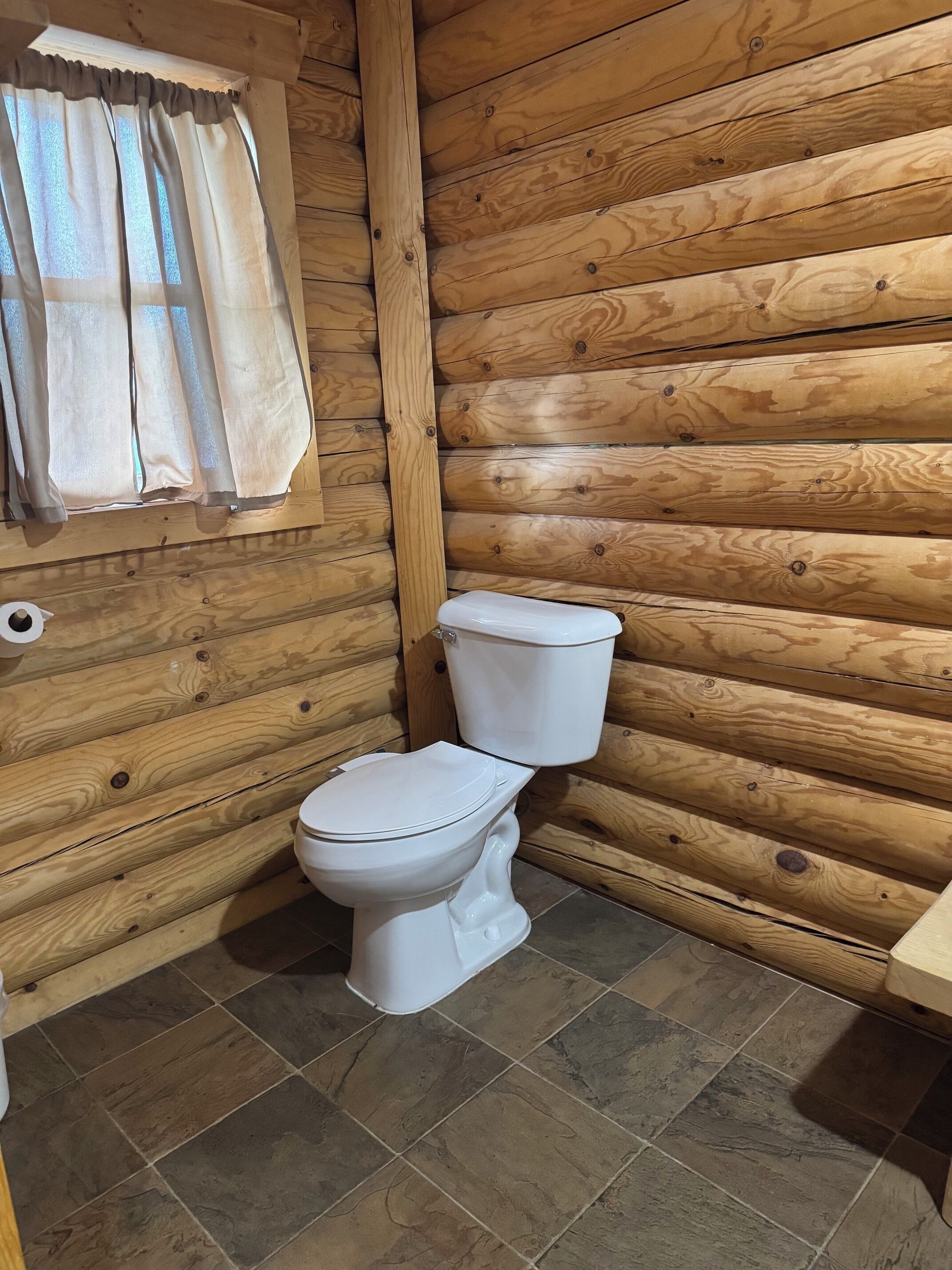Un baño de cabaña de madera con un inodoro, una ventana con cortinas y un rollo de papel higiénico en la pared.